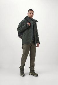 Green insulated jacket, textured gray sweater, olive cargo pants, and green hiking boots. Model carries a gray backpack. Neutral background.