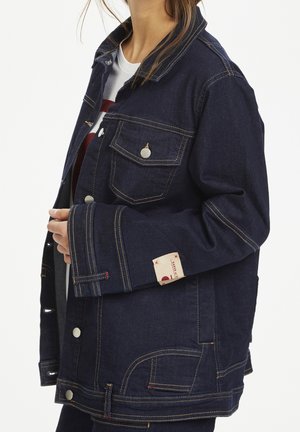 Frau trägt eine dunkelblaue Jeansjacke mit silbernen Knöpfen und sichtbaren gelben Nähten, hält mit einer Hand den Revers der Jacke.