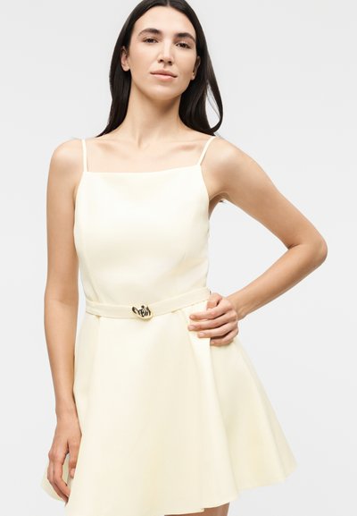 Νεαρή γυναίκα με μακριά σκούρα μαλλιά που φοράει αμάνικο, κρεμ φόρεμα με λεπτές τιράντες και ζώνη στη μέση, πόζαρε με το ένα χέρι στη λεκάνη.