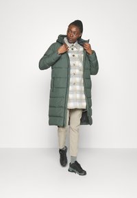 Groene gewatteerde jas met capuchon, voorzien van een ritssluiting en zijzakken, gedragen over een geruite overhemd met knopen, khaki broek en zwarte schoenen.