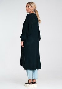 Długi, ciemnozielony sweter z dzianiny o luźnych rękawach, z teksturowanym wzorem i bocznymi rozcięciami, w zestawie z jasnymi dżinsami i trampkami.