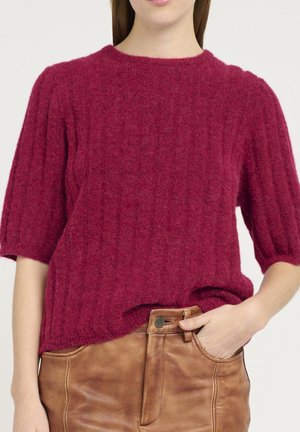Femme portant un pull rouge texturé à manches courtes et un pantalon en cuir marron taille haute, une main dans la poche.