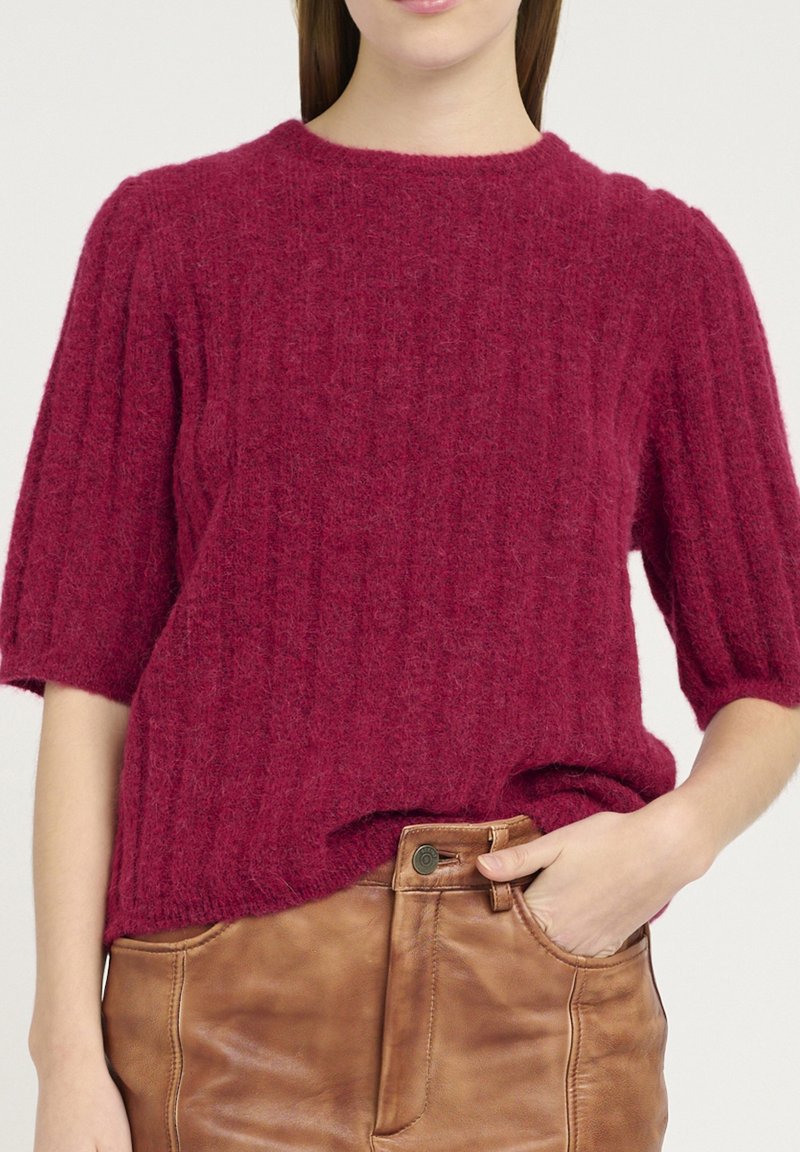 Femme portant un pull rouge texturé à manches courtes et un pantalon en cuir marron taille haute, une main dans la poche.