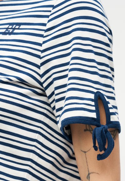 Μπλε ναυτικό και λευκό ριγέ κοντομάνικο πουκάμισο με μικρή σχισμή και κόμπο σε φιόγκο στο μανίκι, που αφήνει να φαίνεται μέρος ενός χρωματιστού τατουάζ στον βραχίονα.