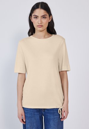 T-shirt beige à manches courtes avec un col rond et un détail de nouage ajustable sur le côté. Fabriqué en tissu doux avec une texture lisse.