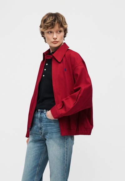 Άτομο με κοντά κυματιστά μαλλιά, φορώντας κόκκινο μπουφάν πάνω από μαύρο πουκάμισο με κουμπιά και ανοιχτό μπλε τζιν, ποζάροντας με ένα χέρι στην τσέπη.