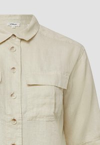 Beige overhemd met korte mouwen, gemaakt van lichte stof, heeft een knoopsluiting aan de voorkant en twee borstzakken. Gladde textuur en klassiek kraagontwerp.