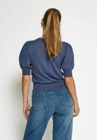 Tamsiai mėlynas megztinis su pūstomis rankovėmis ir raišteliu apačioje, derinamas su aukšto liemens šviesiai mėlynomis džinsomis su užpakalinėmis kišenėmis.