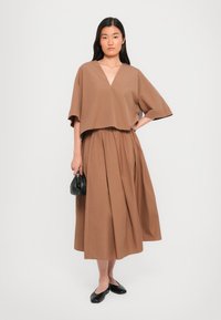Brown, cropped top with a V-neck and wide sleeves paired with a matching, flowy, ankle-length skirt; smooth fabric; black handbag; flat shoes.