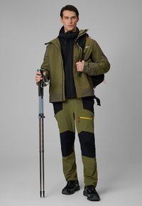 Homme en veste et pantalon de randonnée verts et noirs tenant des bâtons de trekking et portant un sac à dos noir sur fond gris.