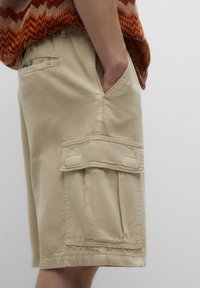 Shorts cargo beiges en coton, dotés de poches latérales, d'une poche à rabat sur la jambe et d'une coupe décontractée avec une texture lisse.