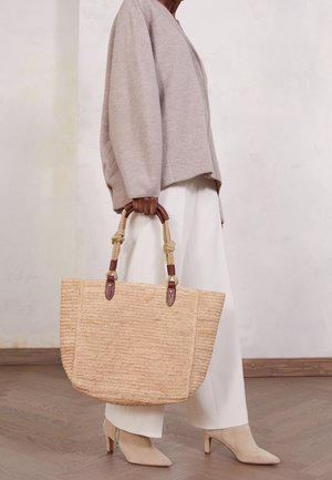 Osoba w beżowym swetrze, białych spodniach i beżowych botkach na obcasie trzyma duży wiklinowy słomiany worek z uchwytami ze sznurka i skóry.