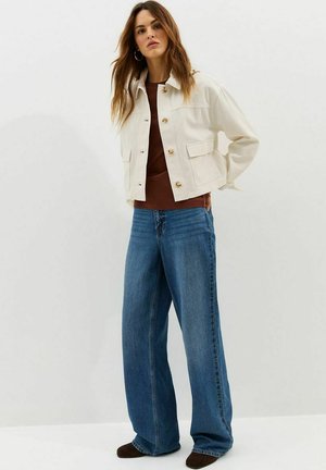 Vrouw staand, gekleed in een crèmekleurig cropped jasje, bruin topje, wijde blauwe jeans en donkere schoenen tegen een effen witte achtergrond.