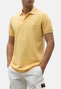 Polo amarillo hecho de algodón texturizado. Presenta un cuello, mangas cortas y un pequeño logo en el lado izquierdo del pecho. Se lleva con pantalones cortos blancos.