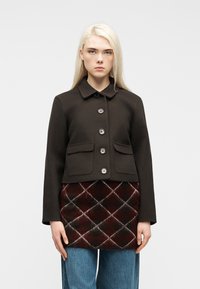 Γυναίκα με μακριά ξανθά μαλλιά φοράει μαύρο σακάκι με κουμπιά πάνω από μια καπιτονέ μπορντό και μαύρη φούστα και μπλε τζιν, στέκεται μπροστά σε λευκό φόντο.