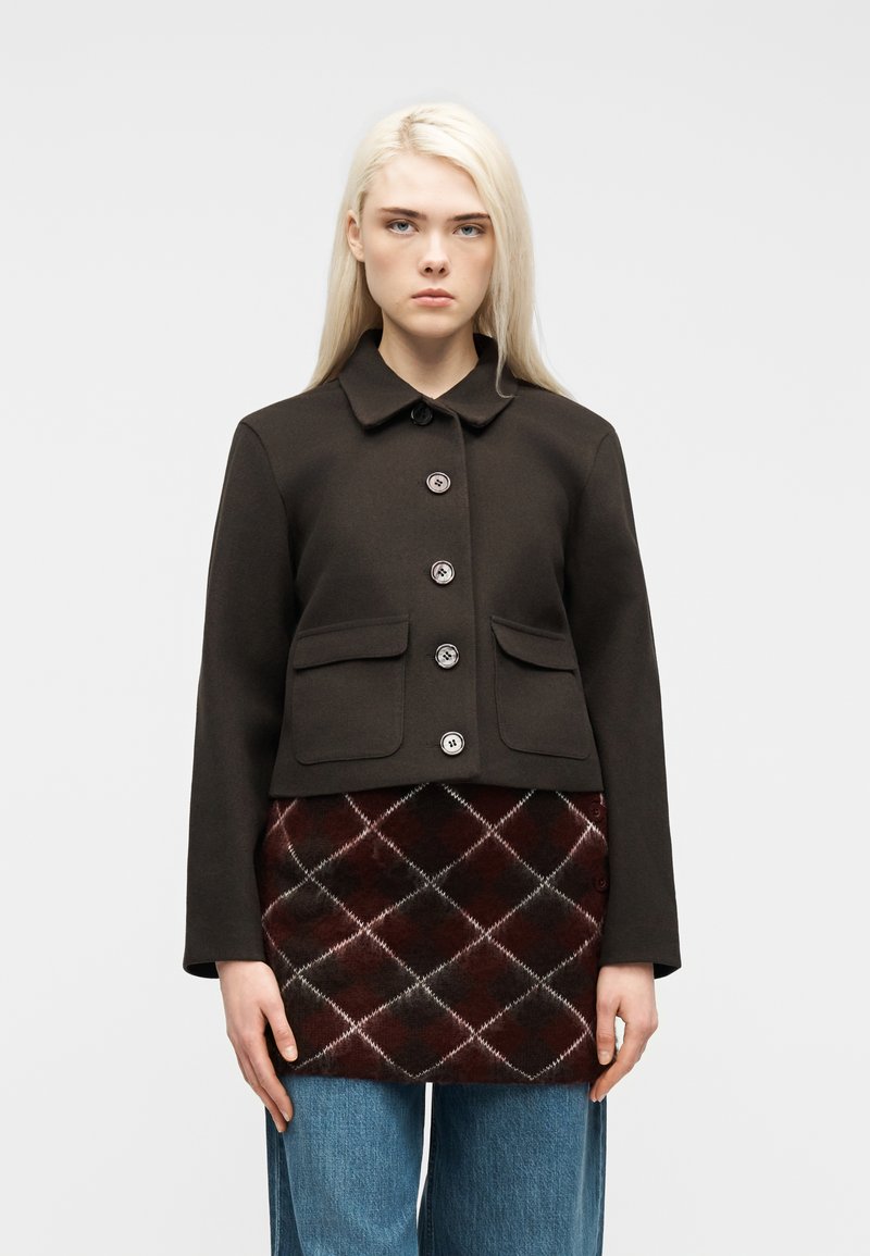 Γυναίκα με μακριά ξανθά μαλλιά φοράει μαύρο σακάκι με κουμπιά πάνω από μια καπιτονέ μπορντό και μαύρη φούστα και μπλε τζιν, στέκεται μπροστά σε λευκό φόντο.