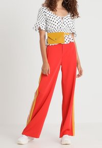 Mujer vistiendo una blusa blanca con lunares, pantalones anchos rojos con rayas amarillas, zapatillas blancas y una riñonera amarilla.
