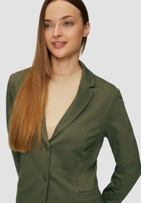 Blazer verde ajustado con solapa con muesca, botones sencillos y tejido texturizado. Llevado sobre un suéter beige claro. Superficie lisa.