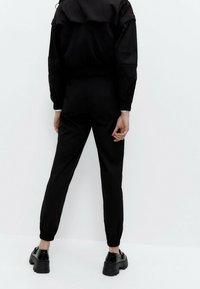 Persoon die naar een witte muur staat, gekleed in zwarte elastische broek met manchet, een zwarte jas en zwarte platform schoenen met kettingdetails.