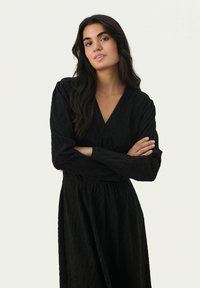 Schwarzes Kleid mit langen Ärmeln und V-Ausschnitt, das mit einem strukturierten Blattmuster verziert ist. Die Taille ist gerafft, was eine fließende Silhouette schafft.