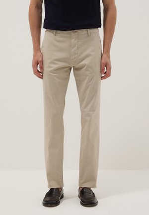 Man draagt beige recht gesneden broek, zwarte loafers en een zwart shirt, staand tegen een eenvoudige lichte achtergrond.