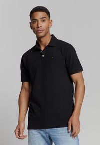 Zwarte poloshirt met een klassieke kraag, korte mouwen, een placket met drie knopen en een klein logo-detail op de borst. Draagt het met lichtblauwe jeans.