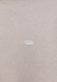 Lichtbeige sweater van een zacht, textuurstof. Een zilveren ovale label met "Simabatik" is op de voorkant gestikt.