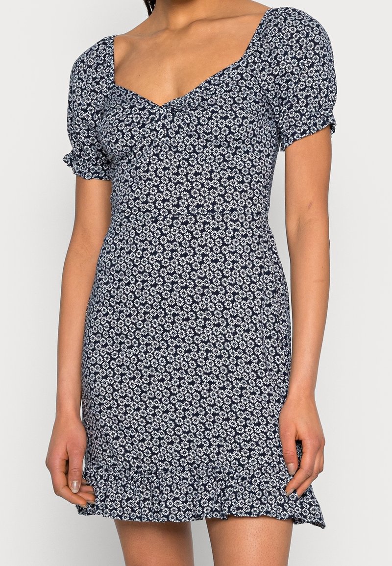 Marinblå klänning med blommönster, korta puffärmar, fyrkantig halsringning och volangkant. Åtsittande design i lättviktsmaterial.