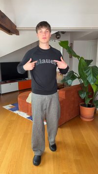 Ung mann står i en stue og peker på ordet "Blauer" på sin svarte sweatshirt, med en stor potteplante og oransje sofa bak.