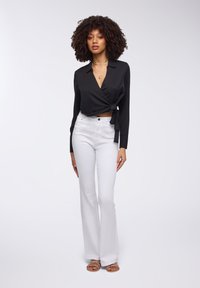 Blusa nera avvolgente con maniche lunghe, legata in vita, abbinata a jeans bianchi a vita alta e a zoccoli piatti. Accenti di gioielleria semplici.