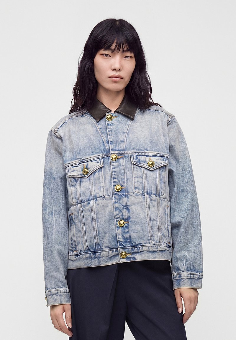 Femme aux longs cheveux noirs porte une veste en jean bleu clair avec des boutons dorés et un col noir, associée à un pantalon foncé, debout sur fond blanc.