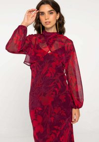 Largo vestido burdeos con mangas de capas translúcidas, con un patrón floral en rojo. Textura suave con una silueta ajustada.
