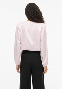 Blouse en satin rose avec manches longues bouffantes et col rond, associée à un pantalon noir taille haute avec une ceinture élastique.