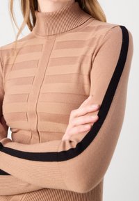 Beige coltrui met ribbelstructuur en horizontale strepen; zwarte accenten die langs de mouwen lopen; aansluitend ontwerp.