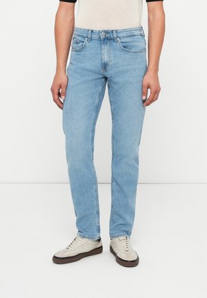 Jeans rectos de mezclilla azul claro con cinco bolsillos, cierre frontal de botón y un desvanecido sutil. Combinados con zapatillas beige.