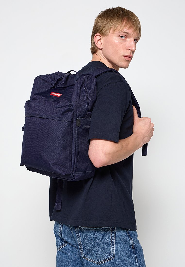 Joven con camiseta negra y vaqueros azules que lleva una mochila Levi's azul marino sobre un hombro, mirando por encima del hombro derecho.