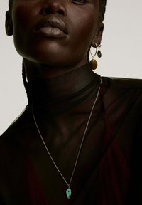 Collar de metal con un colgante de turquesa de diseño intrincado, llevado sobre una blusa negra transparente de cuello alto, acentuado con pendientes dorados.