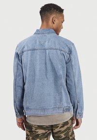 Lichtblauwe denimjas met een klassiek ontwerp. Bevat een kraag, knoopmanchetten en een patch op de bovenrug. Draagt over een tan, grover laagje.