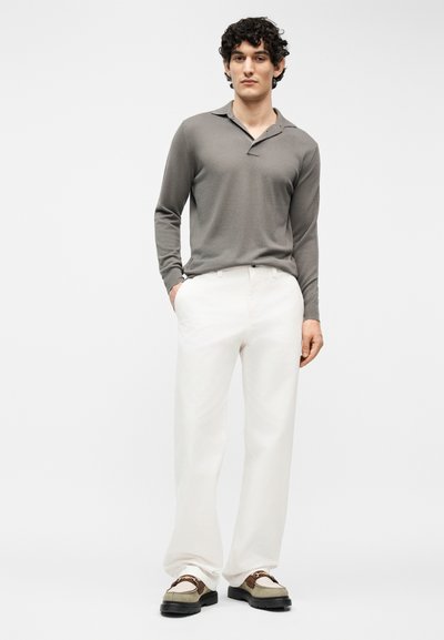 Homme debout les mains dans les poches, portant un polo gris à manches longues, un pantalon blanc et des mocassins beige avec des semelles noires.