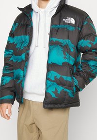 Giacca a vento nera con motivi geometrici turchesi, chiusura con zip e logo sul lato sinistro del petto. Presenta una texture trapuntata e un colletto alto.