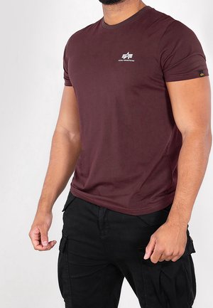 Homme portant un t-shirt ajusté à manches courtes de couleur bordeaux avec un logo blanc sur la poitrine et un pantalon cargo noir, debout devant un fond uni.