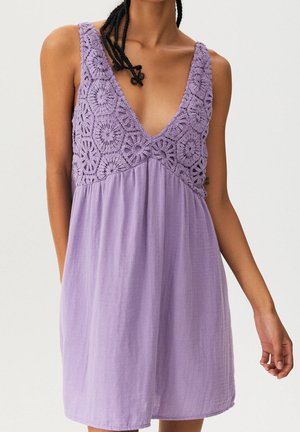 Woman wearing sleeveless lavender dress with deep V-neck and crocheted floral pattern on the bodice.