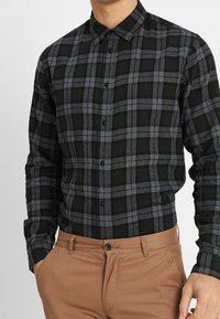 Musta- ja harmaaruudullinen flanellipaita, jossa on napillinen kaulus, pitkät hihat ja suora leikkaus, yhdistetty beigeihin housuihin.