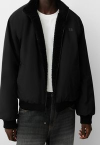 Svart bomberjacka med en fluffig svart krage, framficka med dragkedja och elastiska mudd. Bärs över en vit t-shirt och mörka jeans.