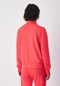 Sudadera roja con cuello alto, mangas largas, puños y dobladillo acanalados, hecha de material suave. Presenta un detalle de logo en el puño.