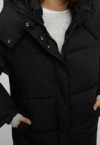 Veste noire matelassée avec fermeture éclair, boutons-pression et capuche doublée. Comprend des poches latérales et des poignets côtelés gris sous les manches.