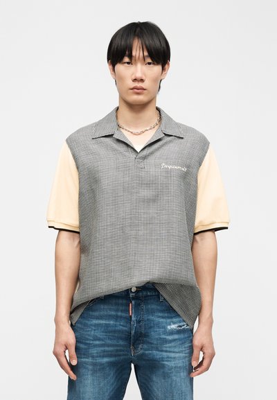 Νεαρός άνδρας με ίσια μαύρα μαλλιά, φοράει μπλούζα με κοντά μανίκια διχρωμία, με μπροστινό μέρος σε ασπρόμαυρο καρό και μανίκια σε ανοιχτό μπεζ χρώμα.