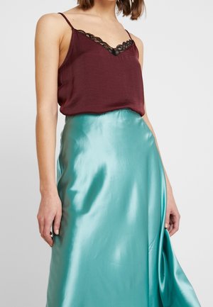 Vrouw die een donkerrode camisole draagt met zwarte kanten rand en een glanzende turquoise satijnen rok, getoond van borst tot midden bovenbeen tegen een effen achtergrond.