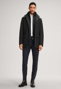 Schwarzer wasserdichter Mantel mit hohem Kragen, Frontreißverschluss und Druckknopfverschlüssen; kombiniert mit schmal geschnittenen, navyblauen Karohosen und schwarzen Lederstiefeln.