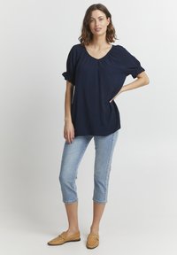 Blusa marina con scollatura a barca, maniche corte a palloncino, abbinata a jeans cropped blu chiaro e mocassini color beige. Tessuto morbido, vestibilità rilassata.